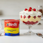 Bird's Original Custard Powder tub with a trifle dessert in a glass bowl on a kitchen counter.